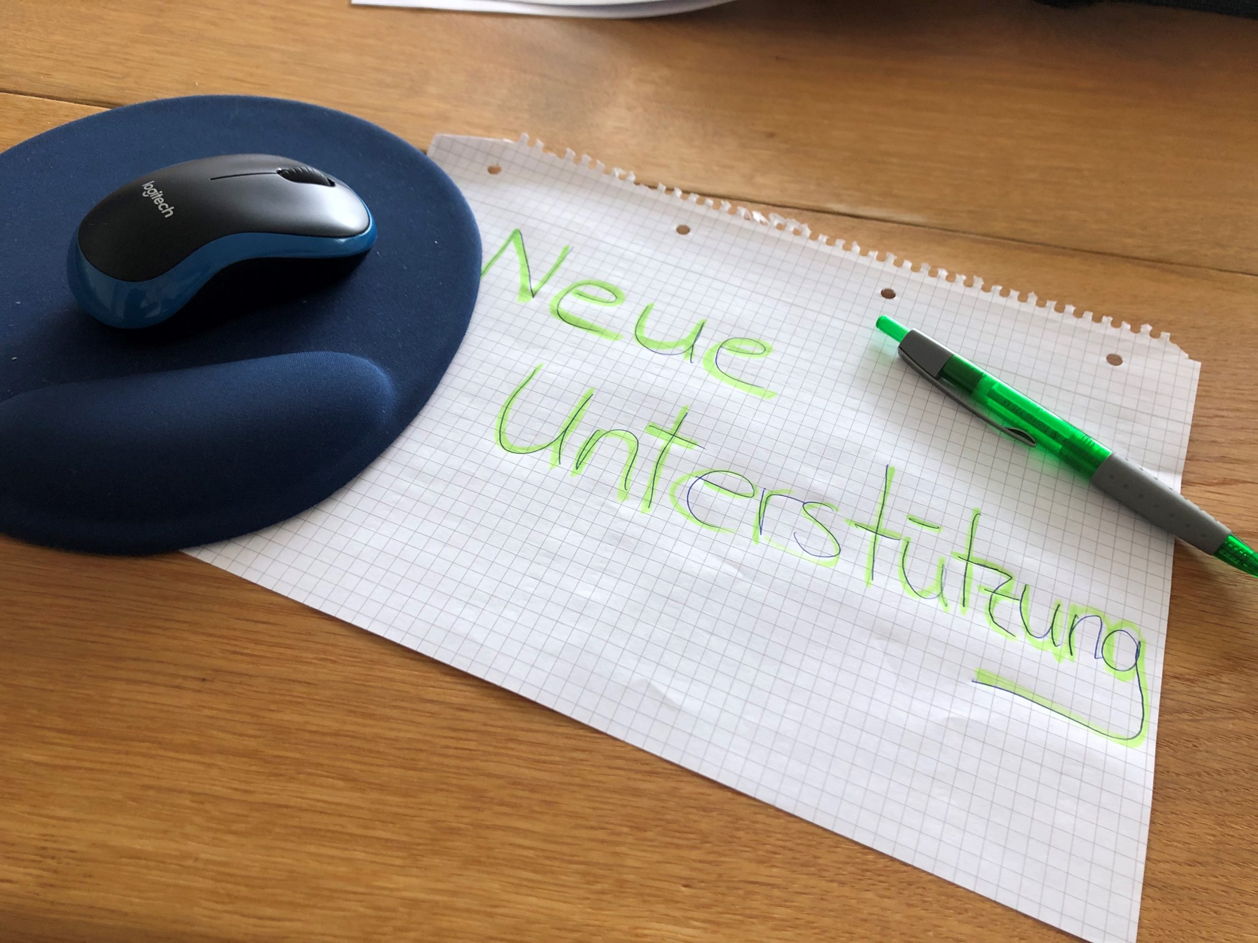 Auf dem Bild ist ein blaues Mousepad zu sehen. Darauf befindet sich eine Computer-Maus. Unter dem Mouspad ist ein kariertes Blatt festgeklemmt. Auf dem Blatt steht: "Neue Unterstützung". Auf dem Blatt liegt oben rechts ein grüner Kugelschreiber.
