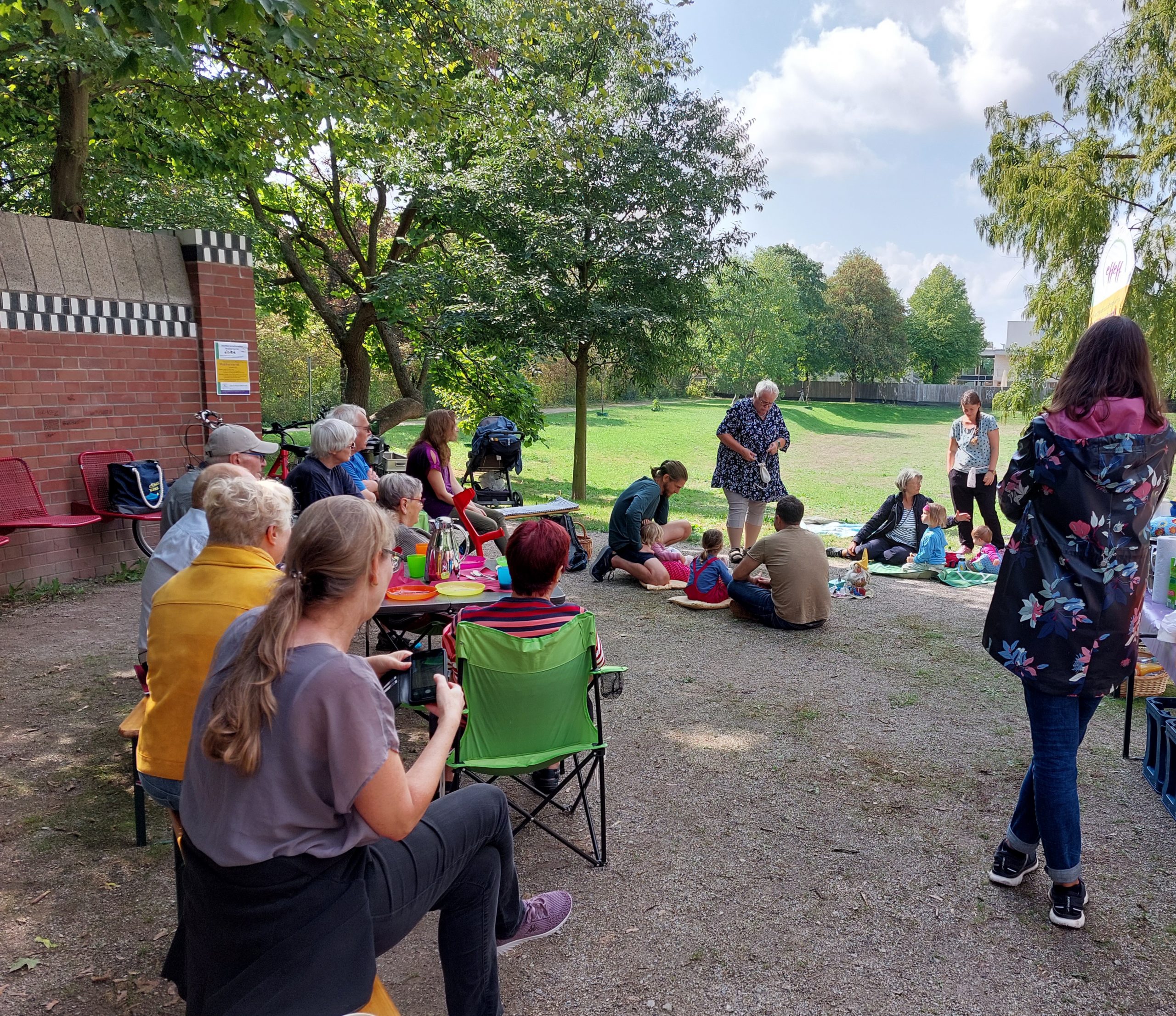 Auf dem Bild ist eine Gruppe von Menschen abgebildet, in deren Mitte eine Märchenerzählerin steht. Die Gruppe trifft sich unter freiem Himmel im Gatschina-Park in Ettlingen.