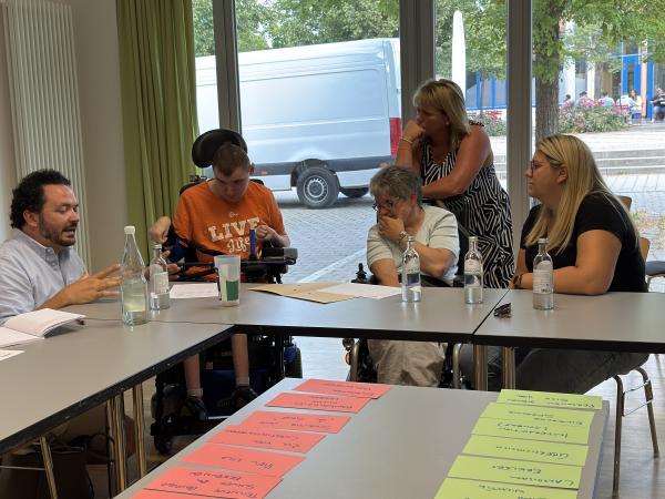 Fünf Menschen an einem Tisch (teils mit Einschränkungen). Eine Person sagt etwas, die anderen hören zu.