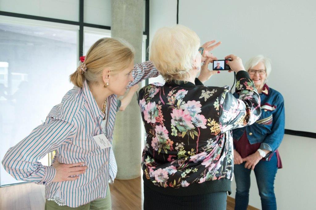 Drei Personen. Zwei Ältere, eine Jüngere. Eine der Älteren wird fotografiert.